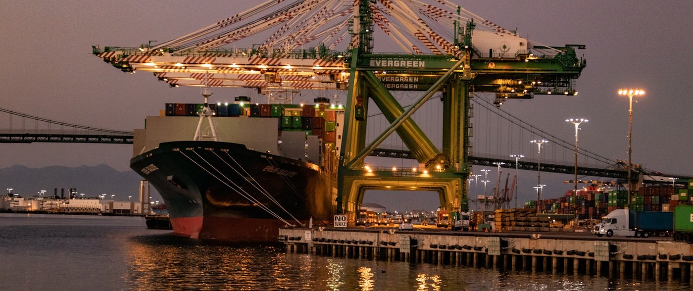 Container ship under a gantry crane at the Port of Los Angeles at dusk, with a truck waiting on the dock